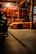A dimly lit garage features a bright orange sports car prominently in the foreground. The garage walls are adorned with several guitars, and a large Budweiser sign hangs above. To the left, part of another vehicle is visible, partially shrouded in shadow.