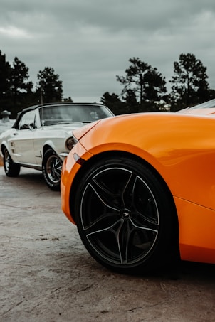 A vibrant orange sports car is prominently displayed in the foreground, showcasing its sleek design and modern wheel with a distinctive pattern. In the background, a classic white car with a vintage aesthetic is visible, parked on a paved surface. Tall, dark trees stand in the backdrop under a cloudy sky, creating an interesting contrast between the modern and classic automotive designs.