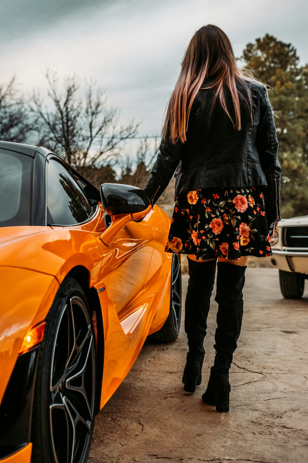 woman in black jacket and black and white floral skirt standing beside orange car