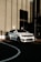 A white SUV is parked beside a tall building featuring large windows and a modern design. The vehicle is positioned on a dark asphalt surface with curved white lines, and long shadows cast by the building indicate a low sun angle. The image suggests an urban setting, highlighting the vehicle's sleek and robust design.