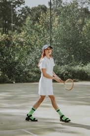 A person is playing tennis on an outdoor court surrounded by greenery. They are wearing a white dress, a dark cap, and distinctive neon green socks paired with black sandals. The tennis racket is wooden, adding a vintage touch.
