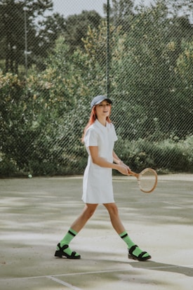 A person is playing tennis on an outdoor court surrounded by greenery. They are wearing a white dress, a dark cap, and distinctive neon green socks paired with black sandals. The tennis racket is wooden, adding a vintage touch.