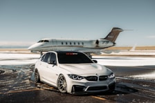 A luxurious car and private jet are parked on a snowy runway. The car, a silver BMW with tinted windows, is in front of the sleek, white aircraft. The setting is an airfield with patches of snow on the ground under a clear sky.