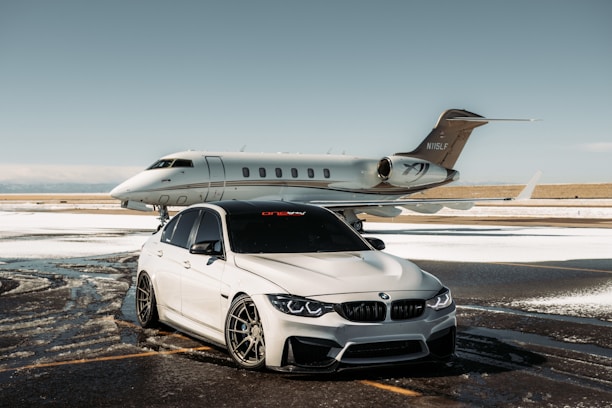 A luxurious car and private jet are parked on a snowy runway. The car, a silver BMW with tinted windows, is in front of the sleek, white aircraft. The setting is an airfield with patches of snow on the ground under a clear sky.