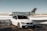 A luxurious car and private jet are parked on a snowy runway. The car, a silver BMW with tinted windows, is in front of the sleek, white aircraft. The setting is an airfield with patches of snow on the ground under a clear sky.