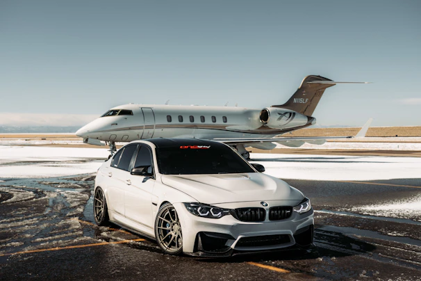 A luxurious car and private jet are parked on a snowy runway. The car, a silver BMW with tinted windows, is in front of the sleek, white aircraft. The setting is an airfield with patches of snow on the ground under a clear sky.