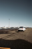 A white car is parked on an expansive asphalt surface near a line of small aircraft. A clear sky provides a backdrop, and the surface reflects some light, indicating recent rain. Tall light poles are visible in the distance.