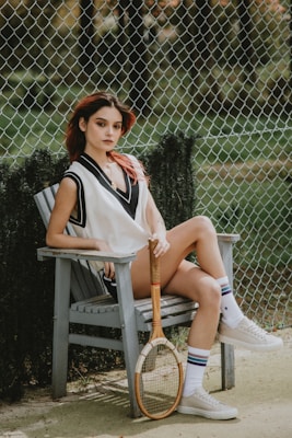 A young person with reddish-brown hair sits on a wooden chair outdoors, holding a wooden tennis racket. They are wearing a sleeveless sports vest, shorts, long socks with stripes, and sneakers. Behind, a chain-link fence and greenery are visible, suggesting a tennis court setting.