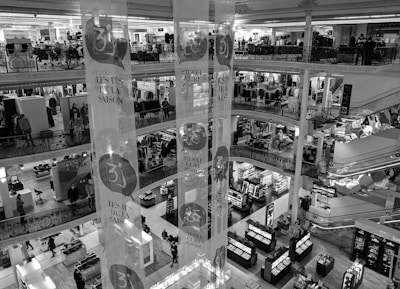 A multi-level department store interior with several floors visible, featuring clothing displays and various sections for shopping. Shoppers are seen walking around, and there are decorative banners hanging from the ceiling. The store has ornate railings and checkered flooring.