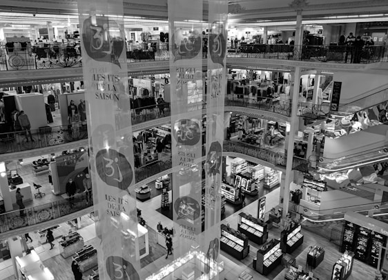 A multi-level department store interior with several floors visible, featuring clothing displays and various sections for shopping. Shoppers are seen walking around, and there are decorative banners hanging from the ceiling. The store has ornate railings and checkered flooring.