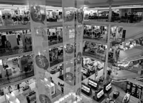 A multi-level department store interior with several floors visible, featuring clothing displays and various sections for shopping. Shoppers are seen walking around, and there are decorative banners hanging from the ceiling. The store has ornate railings and checkered flooring.
