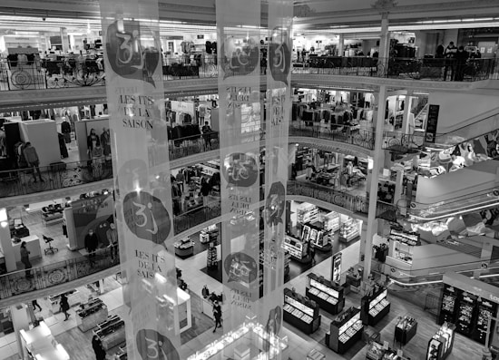 A multi-level department store interior with several floors visible, featuring clothing displays and various sections for shopping. Shoppers are seen walking around, and there are decorative banners hanging from the ceiling. The store has ornate railings and checkered flooring.