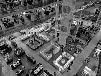 An interior view of a multi-level department store featuring various counters and display units. Mannequins and shelves with items such as clothes, accessories, and other merchandise are visible. The space is well-lit with numerous shoppers moving around.
