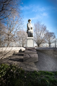 A statue stands prominently on a pedestal in a park setting. The figure is dressed in historical attire and is positioned in front of bare trees, suggesting it is either fall or winter. The background features a clear blue sky, enhancing the overall brightness of the scene. The base of the statue is inscribed with the name 'Penn Treaty Park'. The surrounding area includes greenery and a metal fence.