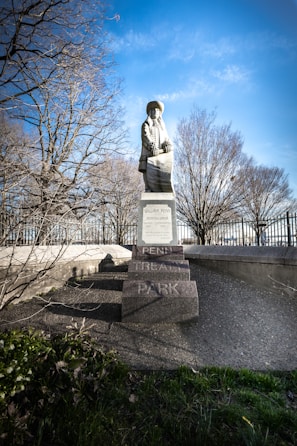 A statue stands prominently on a pedestal in a park setting. The figure is dressed in historical attire and is positioned in front of bare trees, suggesting it is either fall or winter. The background features a clear blue sky, enhancing the overall brightness of the scene. The base of the statue is inscribed with the name 'Penn Treaty Park'. The surrounding area includes greenery and a metal fence.