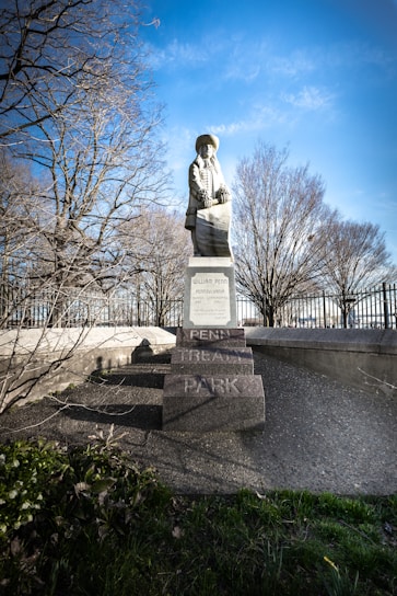 A statue stands prominently on a pedestal in a park setting. The figure is dressed in historical attire and is positioned in front of bare trees, suggesting it is either fall or winter. The background features a clear blue sky, enhancing the overall brightness of the scene. The base of the statue is inscribed with the name 'Penn Treaty Park'. The surrounding area includes greenery and a metal fence.