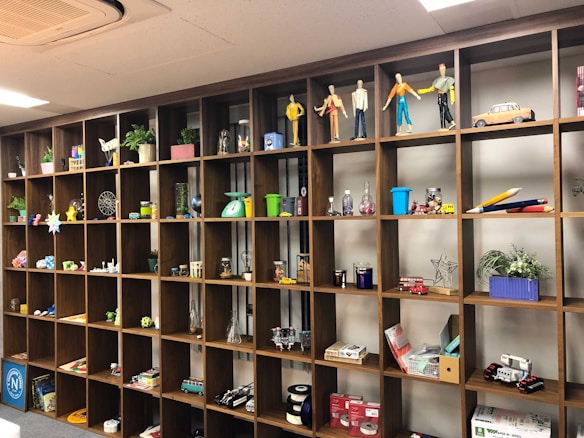 A large wooden shelf with grid-like compartments filled with various objects including figurines, model vehicles, plants, and art supplies. The top section features mannequin-like figures. The shelf is part of a well-lit room with a clean and organized appearance.