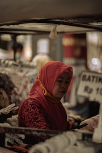 A woman wearing a red hijab and a patterned red top is browsing through racks of clothing in what appears to be a market stall. The focus is on her concentrated expression as she examines the garments closely. Various clothes are hanging from racks, and there is a sign visible in the background with pricing information in Indonesian rupiah.