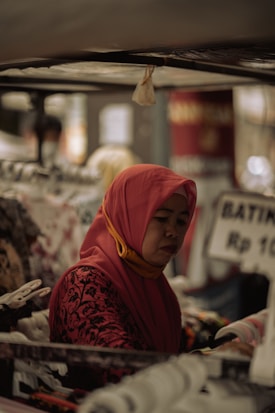 A woman wearing a red hijab and a patterned red top is browsing through racks of clothing in what appears to be a market stall. The focus is on her concentrated expression as she examines the garments closely. Various clothes are hanging from racks, and there is a sign visible in the background with pricing information in Indonesian rupiah.