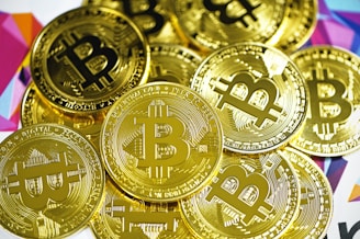 A pile of shiny gold coins featuring the Bitcoin symbol. The coins are closely stacked together and have intricate engraved details. The background includes a colorful geometric pattern with various shades.