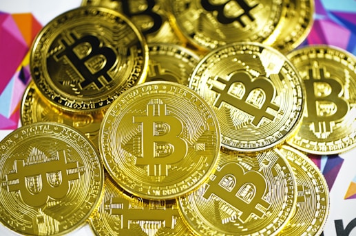 A pile of shiny gold coins featuring the Bitcoin symbol. The coins are closely stacked together and have intricate engraved details. The background includes a colorful geometric pattern with various shades.