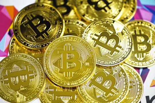 A pile of shiny gold coins featuring the Bitcoin symbol. The coins are closely stacked together and have intricate engraved details. The background includes a colorful geometric pattern with various shades.