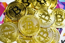 A pile of shiny gold coins featuring the Bitcoin symbol. The coins are closely stacked together and have intricate engraved details. The background includes a colorful geometric pattern with various shades.