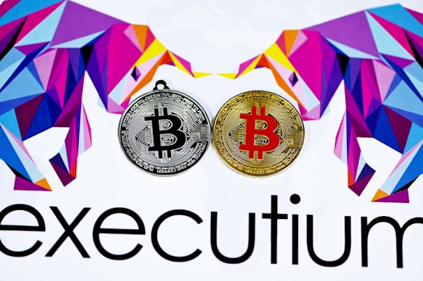 Two stylized Bitcoins, one silver and one gold, are placed side by side. Surrounding them are geometric, colorful designs resembling bulls. The lower part displays the word 'executium'.