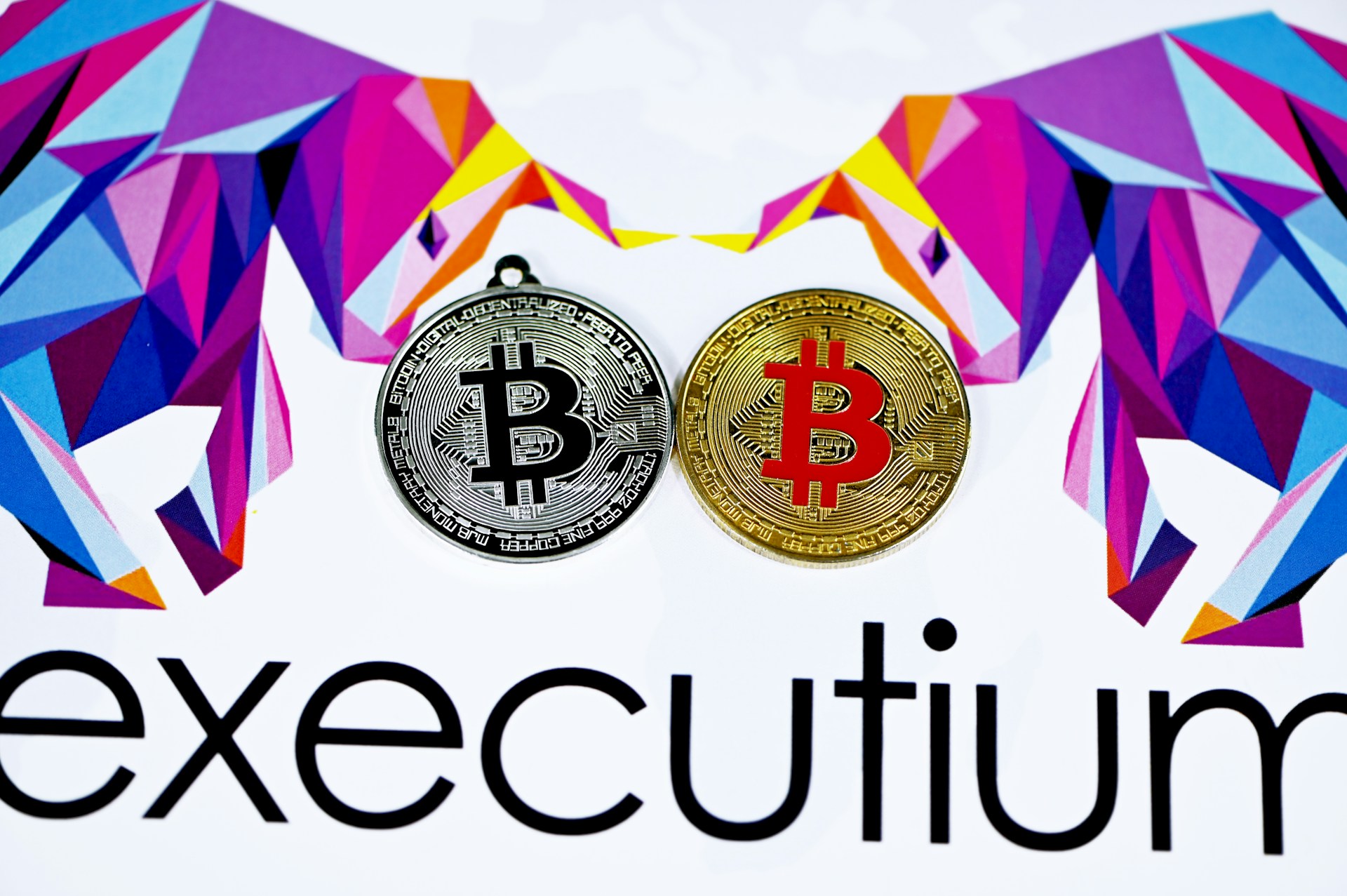 Two stylized Bitcoins, one silver and one gold, are placed side by side. Surrounding them are geometric, colorful designs resembling bulls. The lower part displays the word 'executium'.