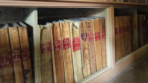 A collection of old, bound volumes of 'The Times' newspaper, dated from the early 1900s, is stored in a wooden and glass display case. The books have a worn, aged appearance with tanned pages and red labels indicating the years.