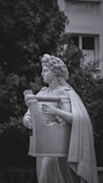 A classical marble statue depicts a figure wearing a laurel wreath and a flowing robe, holding a large lyre. The background features lush greenery and part of a building with a window visible, creating a contrast between the natural and architectural elements.