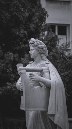 A classical marble statue depicts a figure wearing a laurel wreath and a flowing robe, holding a large lyre. The background features lush greenery and part of a building with a window visible, creating a contrast between the natural and architectural elements.