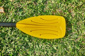 A yellow paddle with a black handle lies on a patch of green grass. The paddle has curved grooves and casts a soft shadow on the grass.