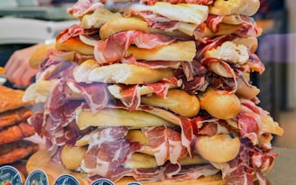 A large stack of assorted sandwiches features slices of ham and crispy baguettes piled high. The sandwiches are artfully arranged, creating a visually appealing and abundant presentation. The scene is colorful and rich in texture, with a focus on the layers of meat and bread.