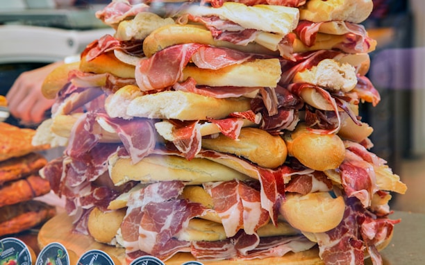A large stack of assorted sandwiches features slices of ham and crispy baguettes piled high. The sandwiches are artfully arranged, creating a visually appealing and abundant presentation. The scene is colorful and rich in texture, with a focus on the layers of meat and bread.