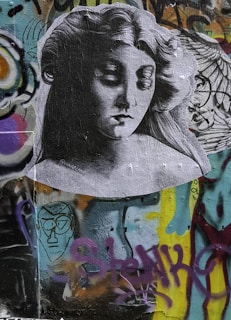 A grayscale image of a woman's serene face is pasted on a colorful graffiti wall. Her expression is calm, with closed eyes and flowing hair. Surrounding her are various graffiti elements, including abstract shapes and spray-painted text in vibrant colors like blue, yellow, and purple.