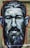 A vibrant street art mural depicting a man's face with detailed expressive features and a beard. The artwork is painted in bold strokes with a mix of colors on a rough textured wall background.