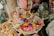 A collection of vibrant flower arrangements placed in shallow baskets made from palm or banana leaves. The flowers include various colors such as pink, yellow, red, purple, and white, set against a background of an eclectic assortment of items including packets on a shelf and stacked biscuits wrapped in plastic.