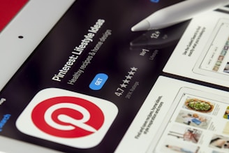 A close-up of a tablet screen displaying the Pinterest app page on a digital store, with the Pinterest logo prominently featured. A white stylus is resting on the screen, which also shows app details like stars rating and install button.
