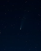 A dark night sky filled with numerous small, distant stars scattered across the image. At the center, a bright object with a tail stretches diagonally, resembling a comet or meteoroid traveling through space. The sky has a deep, almost black hue, which contrasts with the white and slightly blue shades of the celestial bodies.
