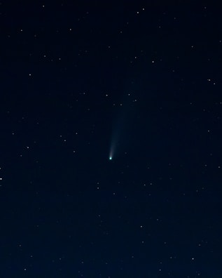 A dark night sky filled with numerous small, distant stars scattered across the image. At the center, a bright object with a tail stretches diagonally, resembling a comet or meteoroid traveling through space. The sky has a deep, almost black hue, which contrasts with the white and slightly blue shades of the celestial bodies.