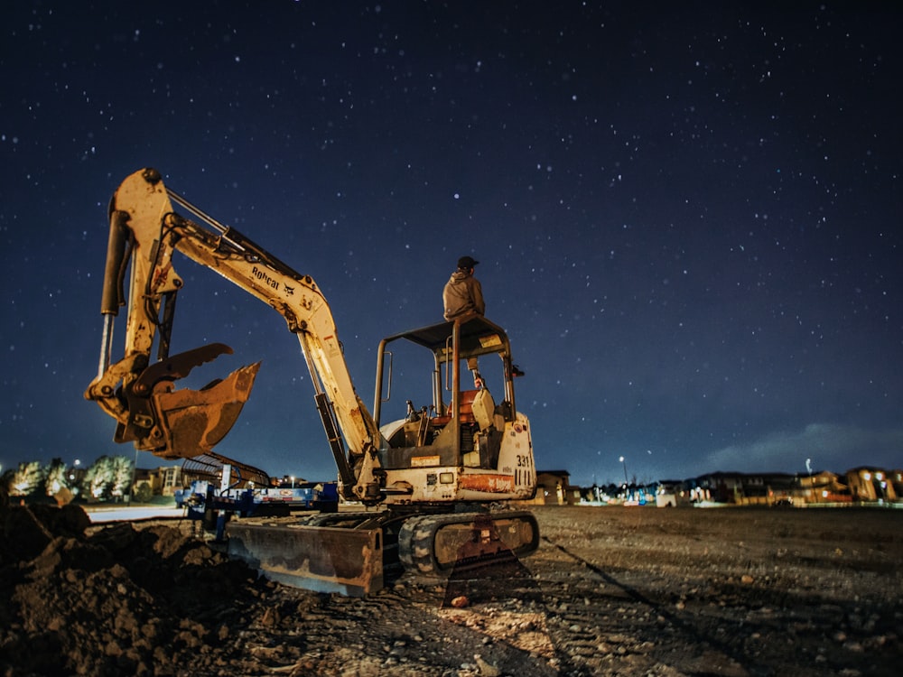 man in brown jacket and blue denim jeans sitting on yellow excavator during night time
