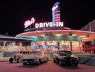 A brightly lit retro diner with neon signs and classic cars parked in front. The sign reads 'Mel's Drive-In' and the exterior features a round structure with large windows showcasing the warm, inviting interior lighting. Classic cars, including a white and a black vintage vehicle, are lined up in front, adding to the nostalgic ambiance.