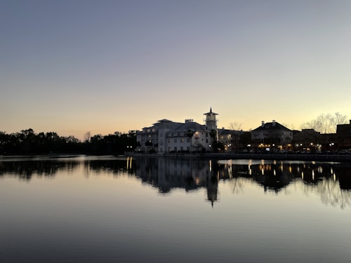 A serene sunset over a tranquil lake, with reflections of buildings and trees on the water's surface. The sky is a gradient of soft blues and oranges, and the silhouette of a small town with a prominent tower stands near the water's edge. Warm lights are illuminated, adding to the peaceful evening atmosphere.