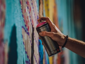 A hand holding a spray paint can with a pink cap, applying paint to a wall covered in colorful patterns. The wall features a variety of vibrant colors including shades of blue, purple, and yellow, creating a textured artistic effect.