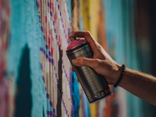 A hand holding a spray paint can with a pink cap, applying paint to a wall covered in colorful patterns. The wall features a variety of vibrant colors including shades of blue, purple, and yellow, creating a textured artistic effect.