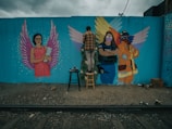 A mural painting on a blue wall features three figures with wings. The left figure is a woman in a pink dress holding a book with purple wings. The center and right figures include a person in a police uniform and a firefighter, both with colorful wings. A person stands on a stool in front of the mural, working on the art.