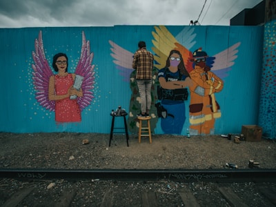 A mural painting on a blue wall features three figures with wings. The left figure is a woman in a pink dress holding a book with purple wings. The center and right figures include a person in a police uniform and a firefighter, both with colorful wings. A person stands on a stool in front of the mural, working on the art.