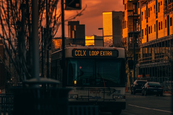 A bus marked with a digital sign 'CCLX Loop Extra' is seen in an urban setting during sunset. The buildings in the background, including skyscrapers, are bathed in a warm orange glow. The street is flanked by bare trees and construction sites. The image evokes a sense of tranquility at dusk.