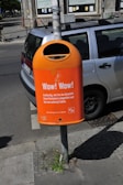 A bright orange public trash bin is mounted on a pole next to a parked car on a city street. The bin has text printed on it, promoting responsible waste disposal, specifically about wrapping and disposing of pet waste. The background shows part of a building with windows and a cyclist in the distance.